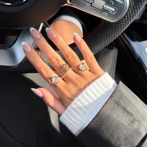 May include: A set of four gold-toned rings worn on fingers, featuring various designs including a rectangular clear stone, a multi-band style, a thin pav&eacute; band, and a teardrop-shaped clear stone, all displayed against a dark steering wheel.