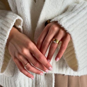 Pode incluir: Close-up de anéis de ouro com um sotaque de diamante. Os anéis são empilhados em um dedo, com uma delicada faixa de ouro em outro dedo. A pessoa está usando um cardigan branco.