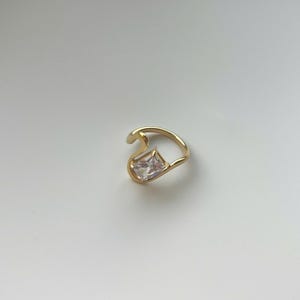 May include: A gold-toned ring featuring an open, asymmetrical band design. The ring is accented with a single, clear, rectangular-cut gemstone set at one end of the curved metal frame, creating a distinct and modern look for the finger.