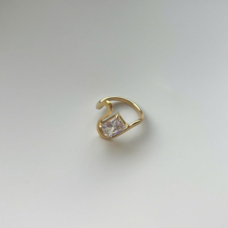 May include: A gold-toned ring featuring an open, asymmetrical band design. The ring is accented with a single, clear, rectangular-cut gemstone set at one end of the curved metal frame, creating a distinct and modern look for the finger.