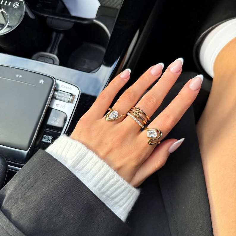 May include: A set of gold-toned rings worn on fingers. One ring features a pear-shaped stone, another has a rectangular stone, and a third is a thin band with small clear stones. The rings are worn alongside a multi-layered gold-toned band ring.