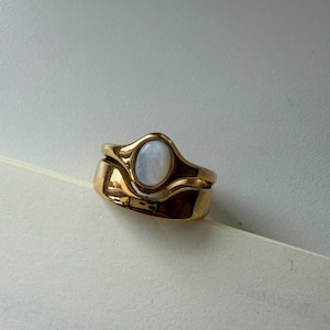 Può includere: Due anelli d'oro impilati. L'anello superiore presenta una pietra ovale di colore chiaro incastonata in una lunetta d'oro. L'anello inferiore è una fascia larga e semplice. Gli anelli sono su una superficie bianca.