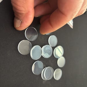 May include: A collection of small, circular, translucent, and clear discs with a smooth, flat surface. These round components are arranged in a loose cluster, featuring a semi-transparent appearance that allows light to pass through each individual piece.
