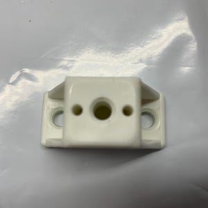 May include: A small, off-white plastic bracket with a rectangular body and two small wings. It has a central hole and two smaller holes on either side, designed for screws or fasteners. The bracket is likely used for mounting or support.