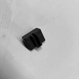 May include: A small, black plastic component with a rectangular shape and two parallel grooves. The object is positioned on a white surface, suggesting a product or part for a larger item.