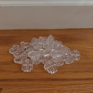May include: A pile of clear, plastic, interlocking building blocks. The blocks are small, with a textured surface and a geometric design. They are scattered on a wooden surface.