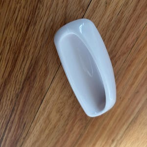 May include: A white, rectangular, ceramic soap dish with a curved interior. The dish is empty and sits on a wooden surface with a natural wood grain pattern.