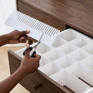 May include: A person is using scissors to cut a white plastic drawer divider. The divider is being placed into a wooden drawer. The drawer is divided into multiple compartments. The drawer is part of a wooden dresser.