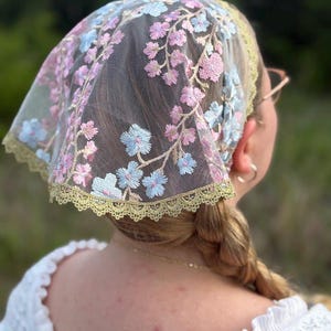Può includere: Un velo trasparente per la testa con dettagli floreali ricamati in rosa e blu. Il bordo è rifinito con un pizzo color oro. Il velo è indossato sui capelli ed è visibile da dietro.