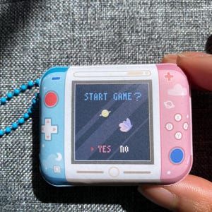 May include: A small, handheld, pink and blue video game console with a screen that says "START GAME?" and has a butterfly and planet graphic. The console has buttons on the side and a blue button on the bottom right corner. The screen also has a "YES" and "no" option.
