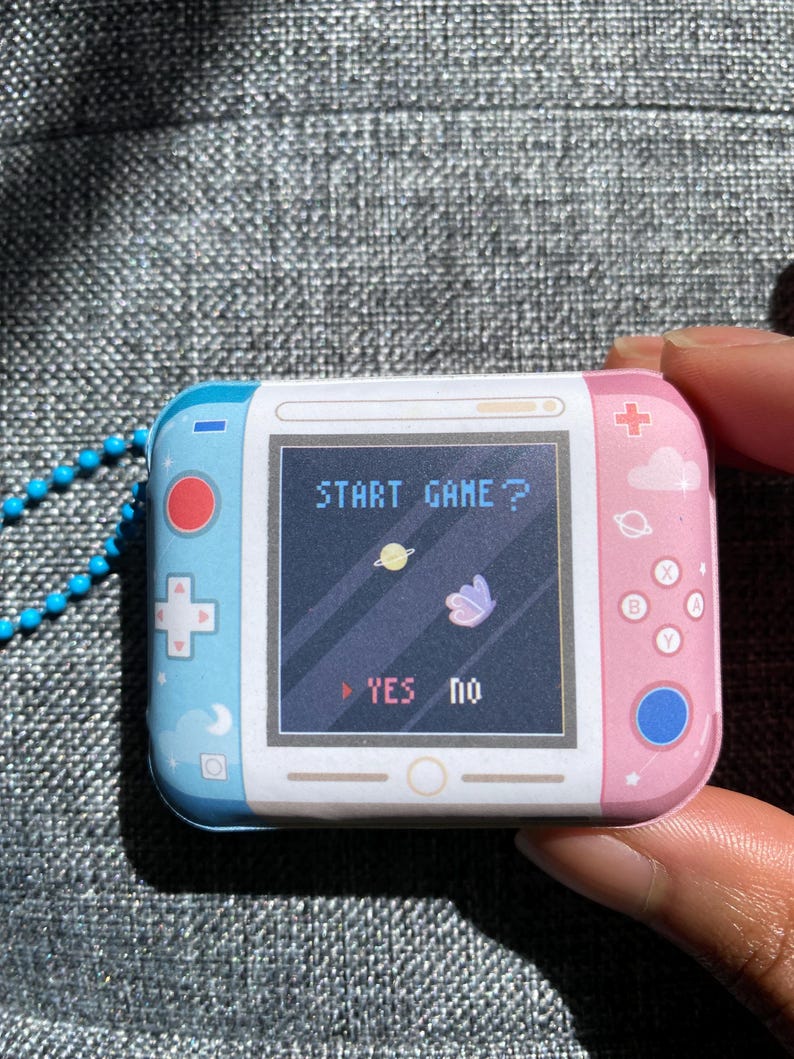 May include: A small, handheld, pink and blue video game console with a screen that says "START GAME?" and has a butterfly and planet graphic. The console has buttons on the side and a blue button on the bottom right corner. The screen also has a "YES" and "no" option.