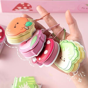 May include: Five colorful, stackable, keychain-sized, fruit-shaped memo pads. The pads are shaped like an orange, an apple, a strawberry, a kiwi, and a broccoli. Each pad has a cute cartoon face and a green leaf.