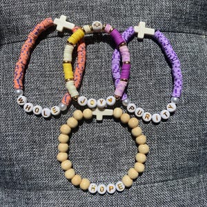 May include: Four bracelets with white cross charms. Three bracelets feature colorful disc beads with the words "WHOLE", "JOY", and "PURITY". The fourth bracelet is made of wooden beads with the word "HOPE".