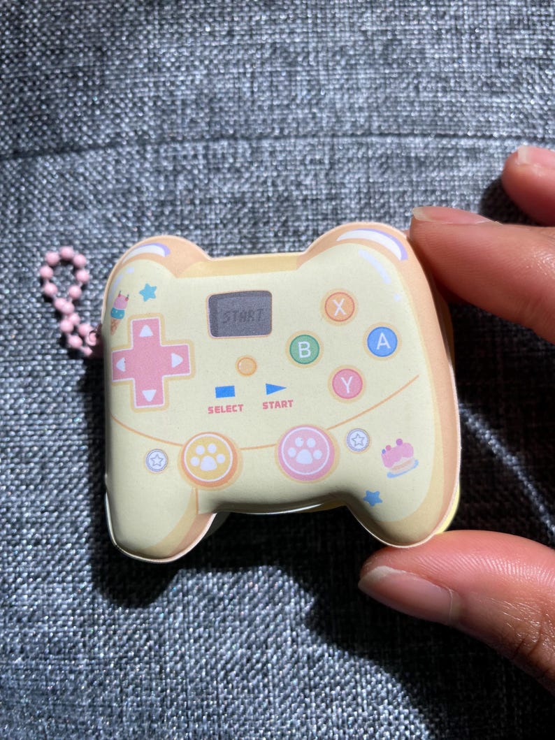 May include: A yellow and white plastic keychain shaped like a video game controller. The controller has pink, blue, and green buttons with the letters X, A, B, and Y. The controller also has a small screen that says "START" and a paw print design.