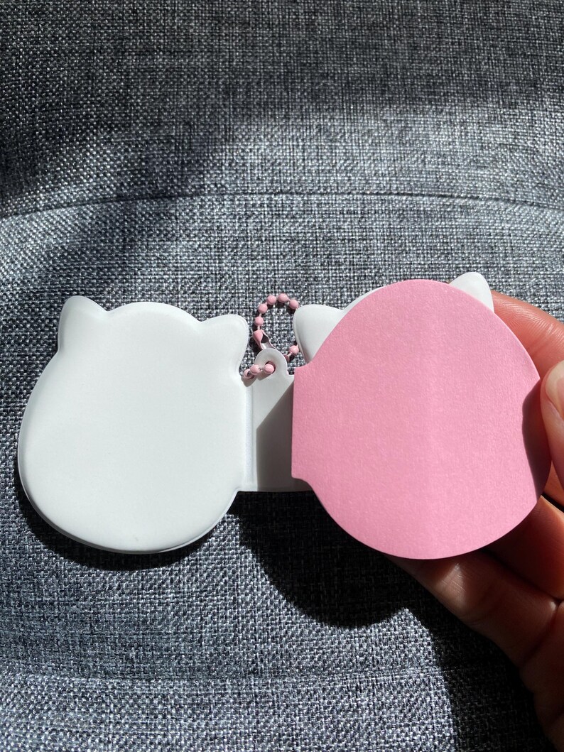 May include: White and pink cat-shaped sticky note holder with a metal chain. The holder has a white front and a pink back.