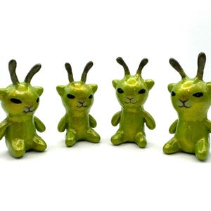 May include: Four small, green, alien-like figurines with gold accents. Each figure has two antennae, a small face, and is sitting with its arms and legs bent. The figurines are arranged in a row against a white background.