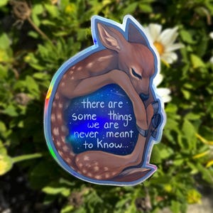 May include: A holographic sticker featuring a fawn curled up with a starry sky inside. The sticker has the text "there are some things we are never meant to know..."