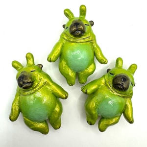 May include: Three whimsical, handmade figurines in a playful pose. Each has a green body with a lighter green belly and a dark brown face. The figurines have a shiny, metallic finish, giving them a unique, artistic look.