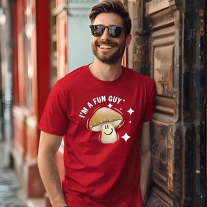 Könnte beinhalten: Rotes T-Shirt mit dem Text "I'M A FUN GUY" und einer Cartoon-Pilz-Grafik. Der Pilz hat ein lächelndes Gesicht und ist von weißen Sternen umgeben. Das Shirt ist kurzärmelig.