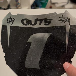 May include: Black and silver sticker with the word "GUTS" and the number "1" in a bold font. The sticker has a glittery texture and the Stussy logo. The text "TMGDECALS" is at the bottom.