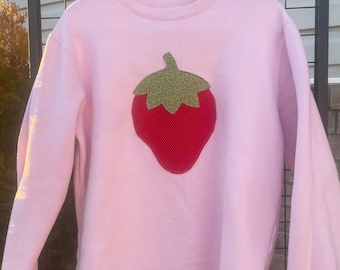 May include: A pale pink sweatshirt featuring a large strawberry design. The strawberry has a red body and a green textured top. The sweatshirt is suspended from a wooden hanger.