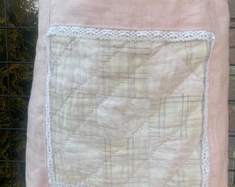 May include: A light pink tote bag with a quilted front panel. The panel is cream-colored with a plaid pattern and trimmed with white lace. The bag has matching pink handles and is made of a soft fabric.