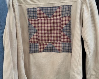 May include: A beige corduroy button-down shirt featuring a large square patch on the back. The patch displays a star pattern created from blue and burgundy checkered fabric. The shirt has long sleeves with buttoned cuffs and a standard collar.