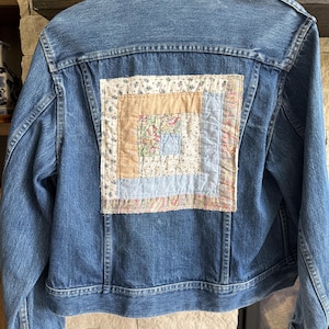 May include: A blue denim jacket featuring a square patchwork panel sewn onto the back. The panel displays a log cabin quilt pattern with various fabric squares in shades of light blue, beige, and floral prints with pink and green accents.