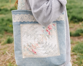 May include: A denim tote bag with a floral patchwork design. The bag features a square patch with pink and white floral patterns, framed by delicate lace trim. The bag is light blue and has a matching lace trim along the top edge.