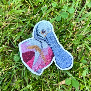 May include: Embroidered sticker of a roseate spoonbill bird. The sticker features a blue head and bill, pink and maroon wing feathers, and a white border. The sticker is placed on a bed of green grass.