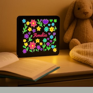 May include: A black, square night light featuring a floral design in pink, yellow, purple, and blue, with the name "Amelia". The night light is on a wooden surface next to an open book and a stuffed bunny. The light is glowing.