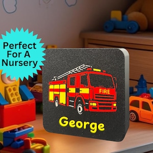 May include: A black square night light with a red and yellow fire engine design and the name "George" in yellow. The word "FIRE" is visible on the truck. The night light is surrounded by children's toys.