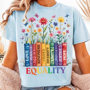 May include: Light blue t-shirt with a graphic of colorful flowers growing from a row of books. The books have words printed on their spines, including "Love", "Peace", "Inclusion", "Kindness", "Chance", "Diversity", "Unity", "Compassion", "Justice", and "Hope". The words "EQUALITY" are printed below the books in rainbow colors.