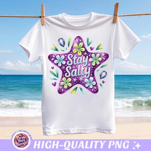 以下が含まれることがあります： 白いTシャツ。紫色のヒトデのデザインで、花と葉で飾られ、「Stay Salty」の文字が入っています。Tシャツは、ビーチと海の背景を背景に、物干しロープに吊るされています。