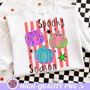 May include: A white long-sleeve sweatshirt featuring a graphic print of three pumpkins in purple, green, and teal with gold speckles. The design includes vertical coral stripes, black star accents, and the text Spooky Season in black lettering.