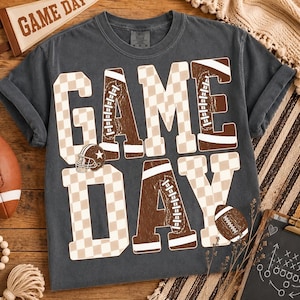 Puede incluir: Una camiseta de manga corta gris carbón con el texto GAME DAY en letras grandes. Las letras tienen un patrón de cuadros beige y blanco, con texturas de balón de fútbol americano integradas en el diseño y un pequeño gráfico de casco.