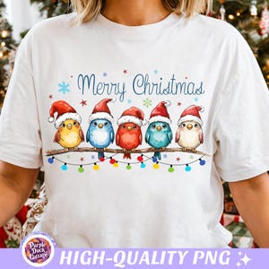 May include: White t-shirt featuring a whimsical Christmas design. The graphic includes five colorful birds wearing Santa hats perched on a branch, with the words "Merry Christmas" above them. Colorful Christmas lights are strung below the birds.