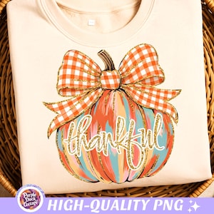 May include: A cream-colored t-shirt featuring a graphic of a pumpkin with orange and white checkered bow. The pumpkin has vertical stripes of orange, blue, and pink with gold glitter accents. The word thankful is written in gold cursive script.
