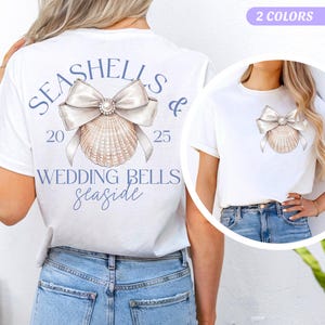 May include: White t-shirt with the text "Seashells & Wedding Bells Seaside" and a seashell with a pearl and a white bow on the back. The front of the shirt has a seashell with a pearl and a white bow.