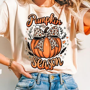 May include: A light beige t-shirt with a graphic of an orange pumpkin with a leopard print bow. The text "Pumpkin Season" is printed in black below the pumpkin.