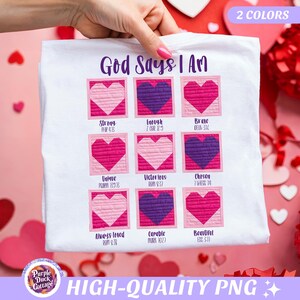 Puede incluir: Camiseta blanca con el texto "God Says I Am" y diseños de corazones coloridos. Cada corazón tiene una palabra como "Fuerte" o "Valiente" y una referencia bíblica. La camiseta es sostenida por una persona.