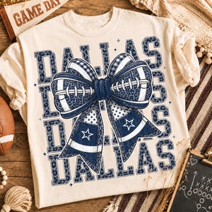 Puede incluir: Una camiseta color crema con la palabra DALLAS repetida en un patrón de leopardo azul oscuro. En el centro, un gran lazo decorativo presenta una textura de balón de fútbol, detalles de cuadros azules y blancos y dos cintas azules con estrellas blancas.