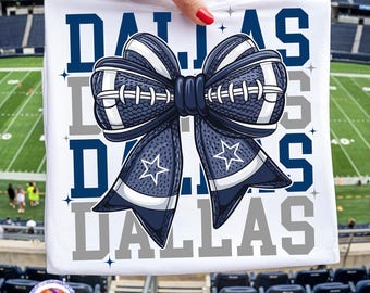 May include: A white t-shirt featuring a large graphic of a navy blue and white bow designed with a football texture and white laces. The word DALLAS is printed in large navy blue and grey block letters behind the bow, accented by small stars.