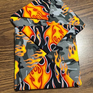 May include: A folded fabric item featuring a flame design in orange, yellow, and black, contrasted with a gray camouflage pattern. The item is likely a coaster or small decorative piece.