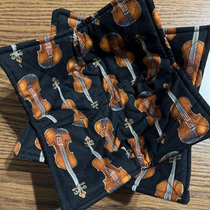 May include: Black quilted pot holders with a repeating pattern of brown violins. The pot holders are star-shaped and have a contrasting black border. The violins have detailed shading and are arranged in various orientations.