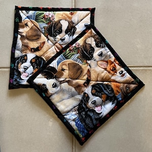 May include: Two square quilted pot holders featuring a colorful fabric print of various puppy breeds including golden retrievers, Bernese mountain dogs, and corgis. The pot holders have a black fabric border decorated with small multicolored paw prints.