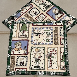 May include: Two square quilted potholders featuring a patchwork print of snowmen, Christmas trees, and candy canes on a cream background. The fabric includes the text "noel", "peace", and "love". The edges are finished with a dark green plaid trim.