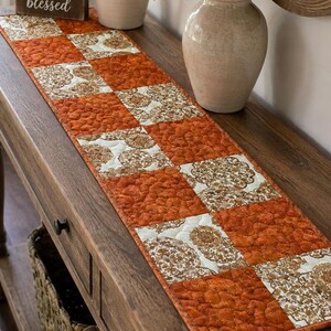 May include: A rectangular quilted table runner featuring a repeating checkerboard pattern of solid burnt orange fabric and cream fabric with a brown floral print. The textile has visible stitching throughout and is laid out flat.