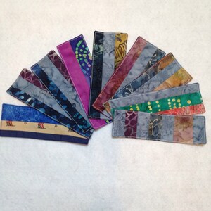 May include: A collection of 12 handmade fabric bookmarks with a variety of colorful patterns and designs. The bookmarks are all rectangular and have a white border.