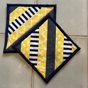 May include: Two square fabric pot holders featuring a patchwork design of yellow and white polka dots, navy blue and white stripes, yellow and white quatrefoil patterns, and navy blue fabric with tiny white dots, all finished with a navy blue border.